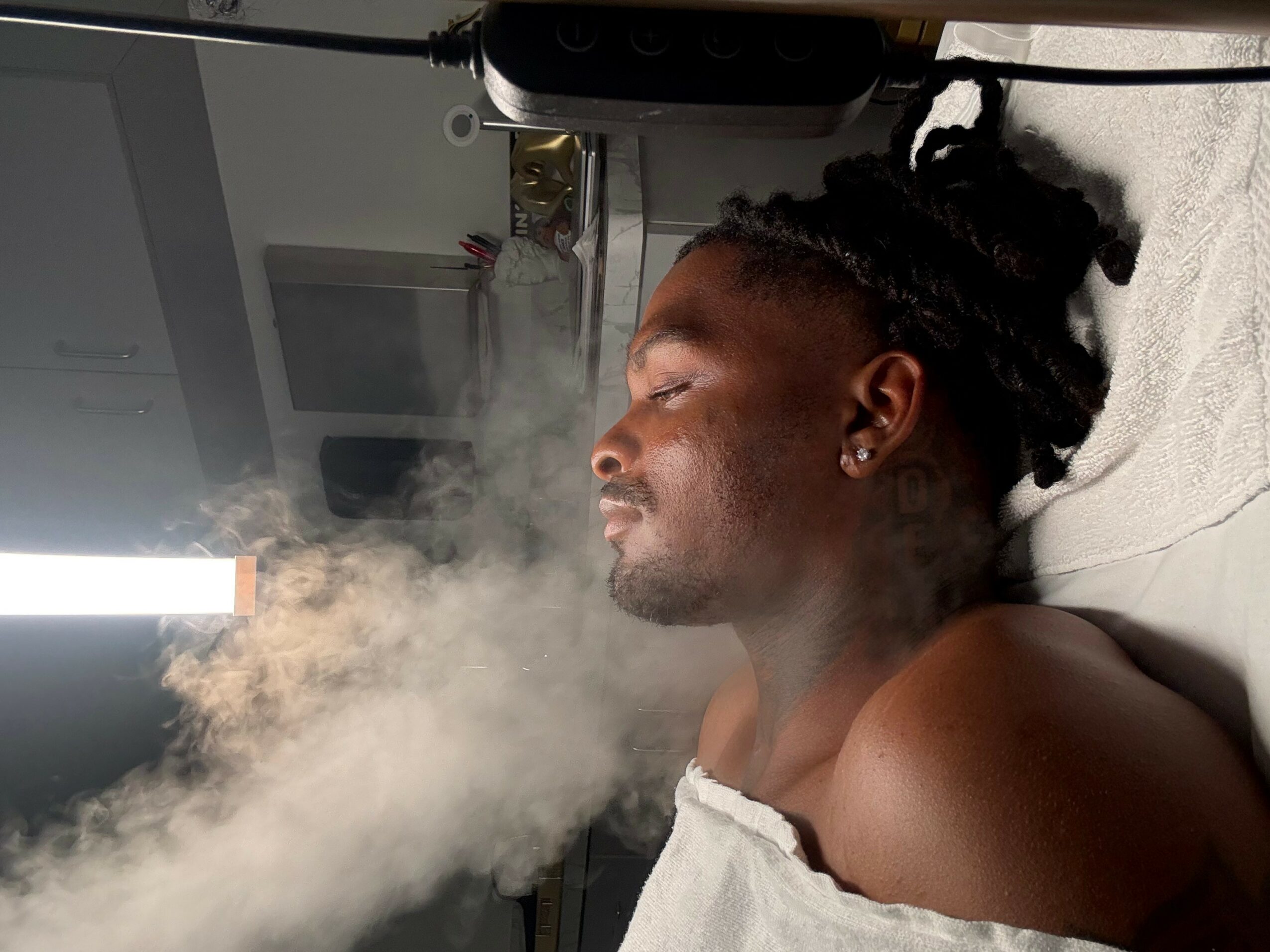 Person lying down with eyes closed, exhaling vapor or smoke, in a room with a window and electronic equipment.