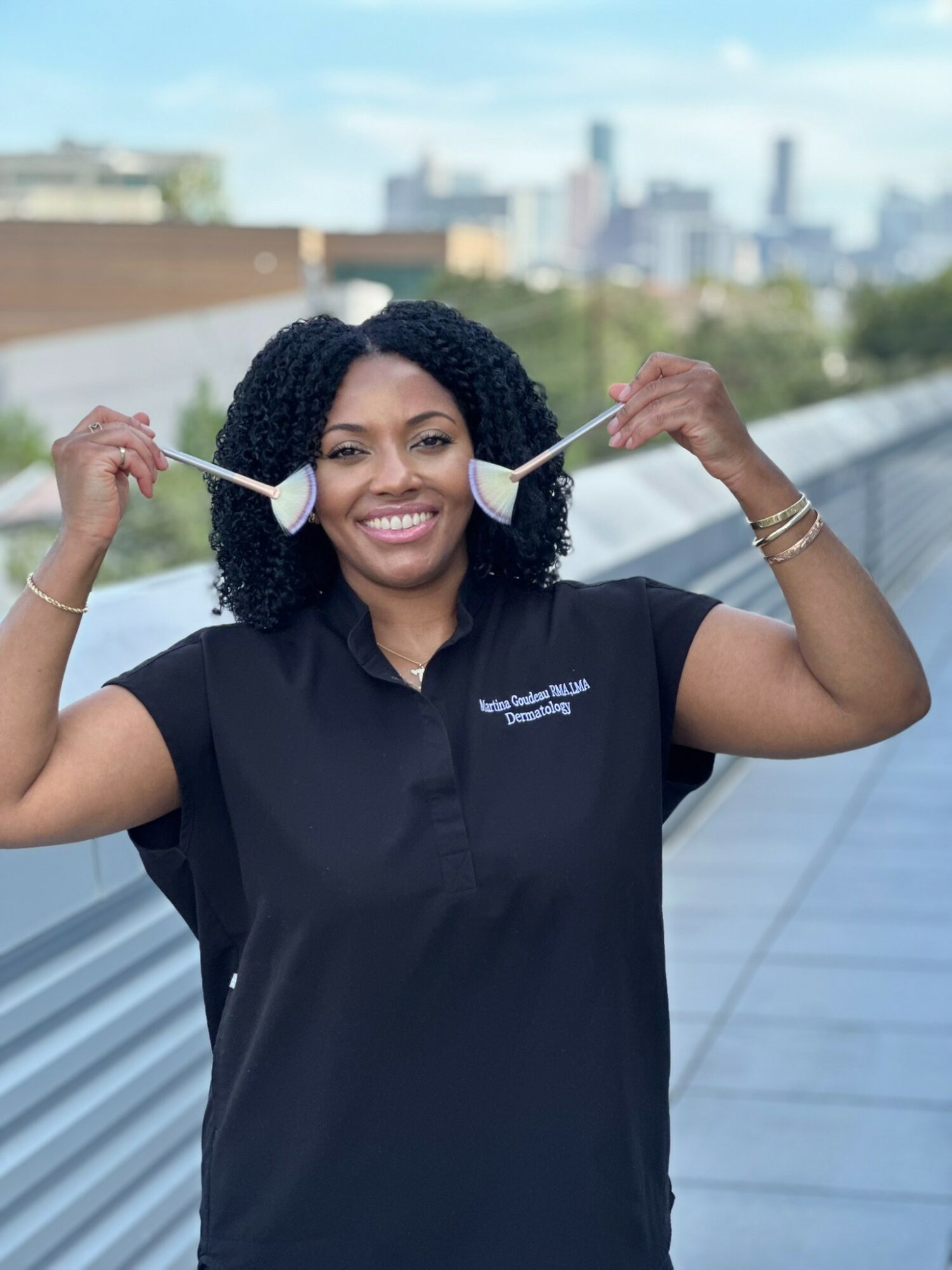 Smiling woman with curly hair holding two face masks outdoors, city skyline in background.