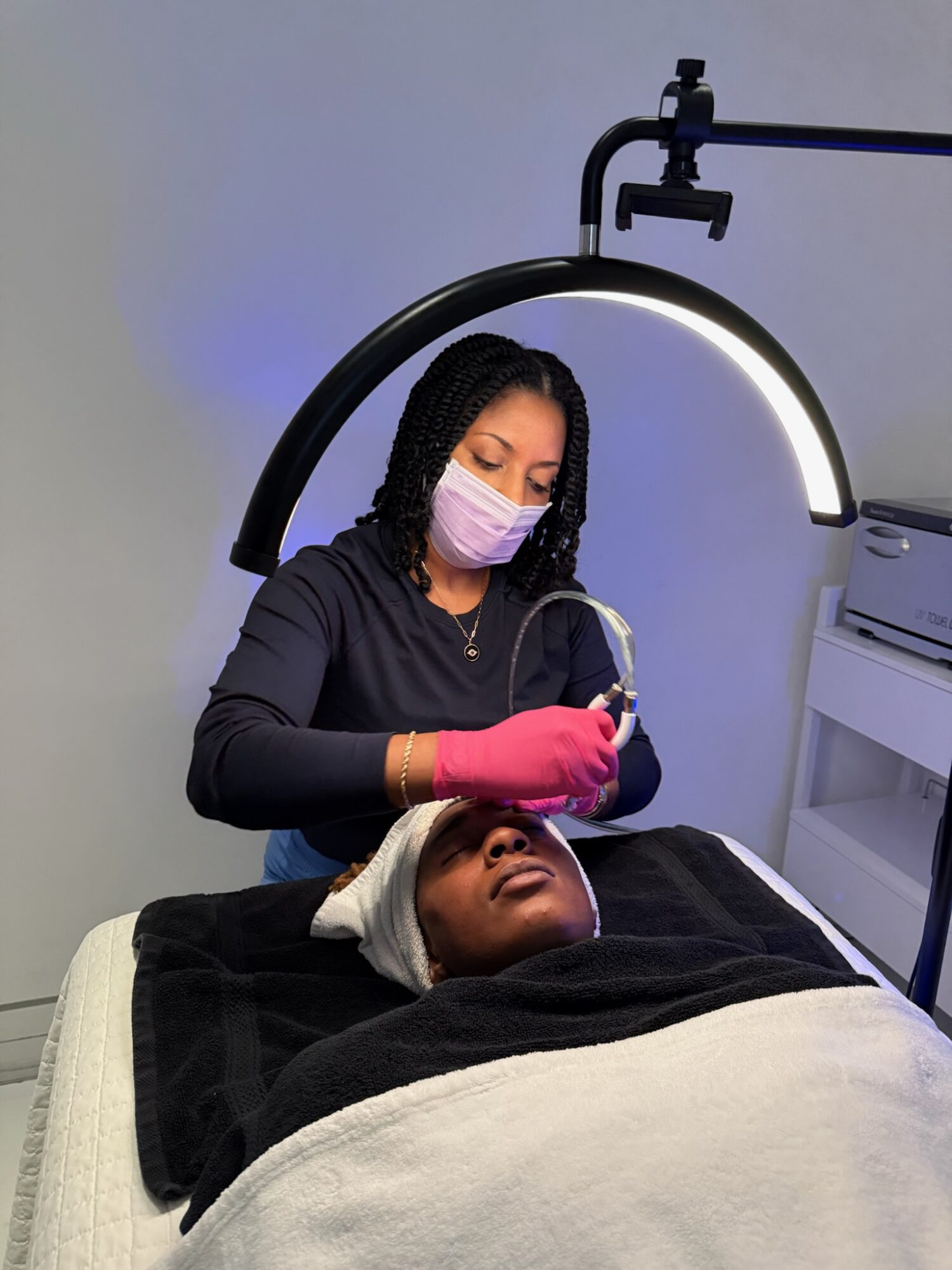 A woman in pink gloves and a face mask performs a procedure on a person's face under a circular light.