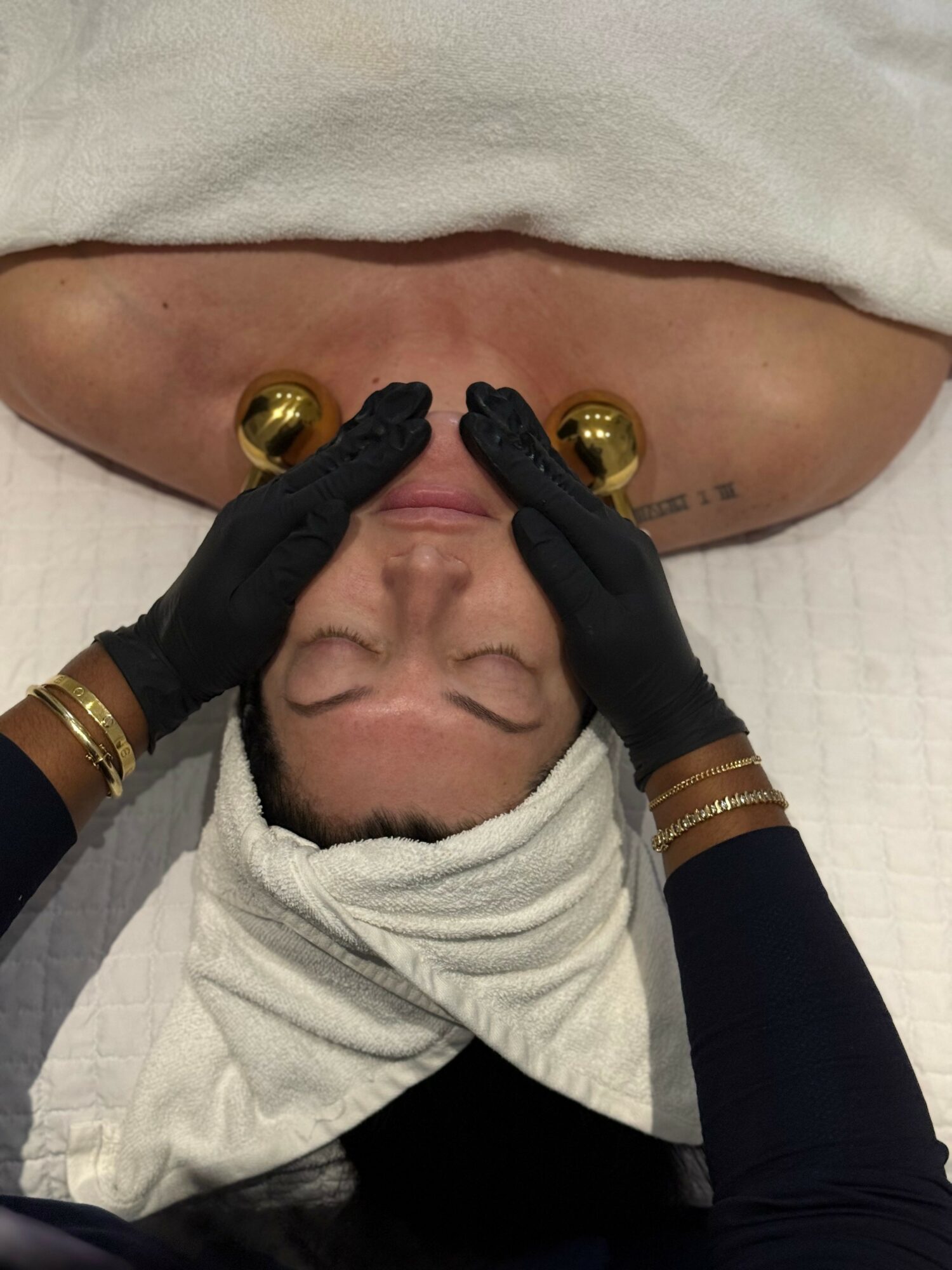 Person receiving facial treatment with hands wearing black gloves, lying down with head wrapped in a towel, two gold massage balls on face