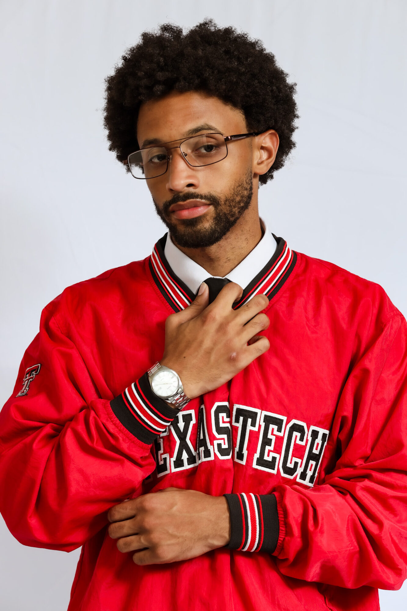 Man with glasses and curly hair adjusting his red jacket with Texas Tech logo, standing against a plain background.