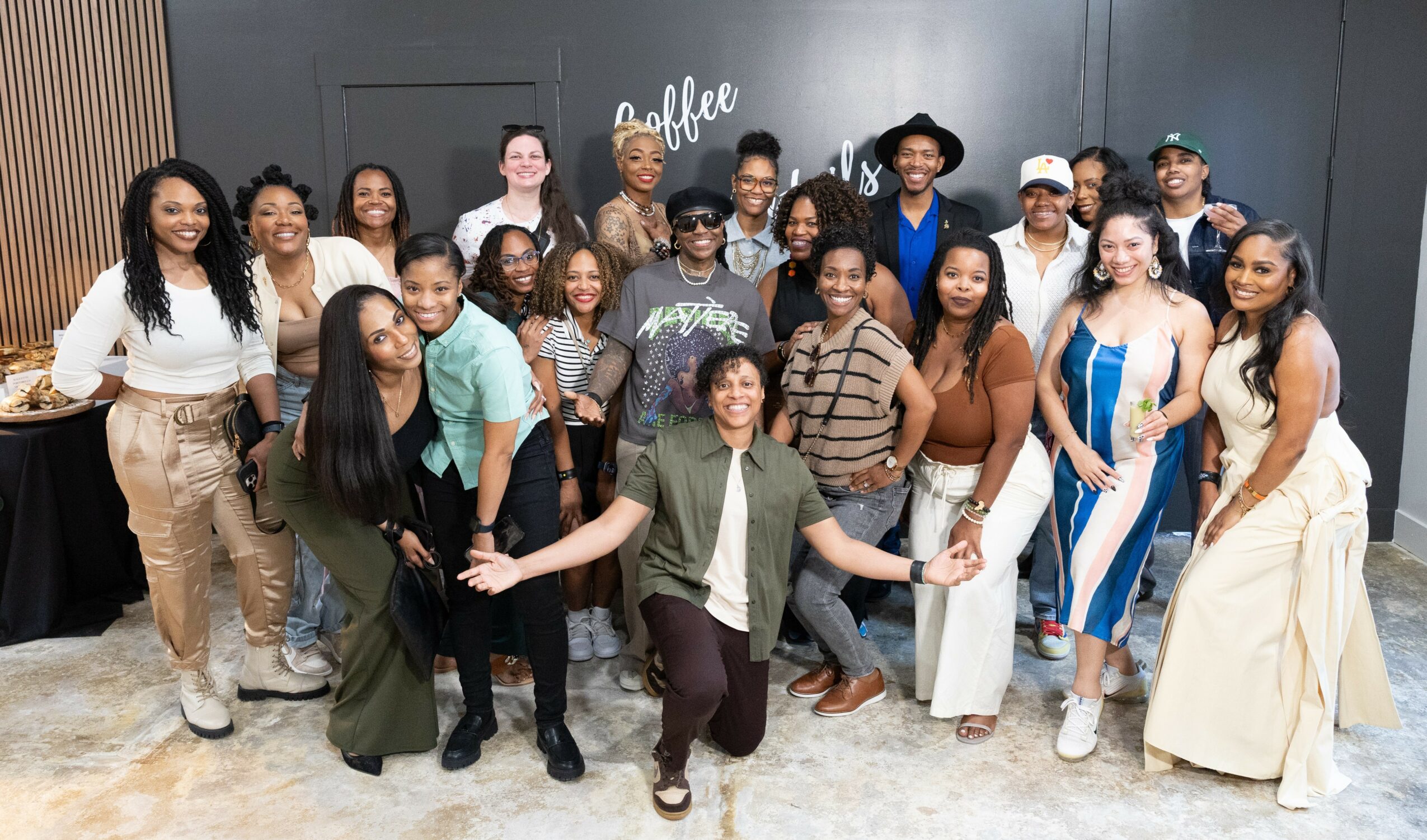 Group of diverse people smiling and posing indoors in front of a black wall with coffee-themed writing.