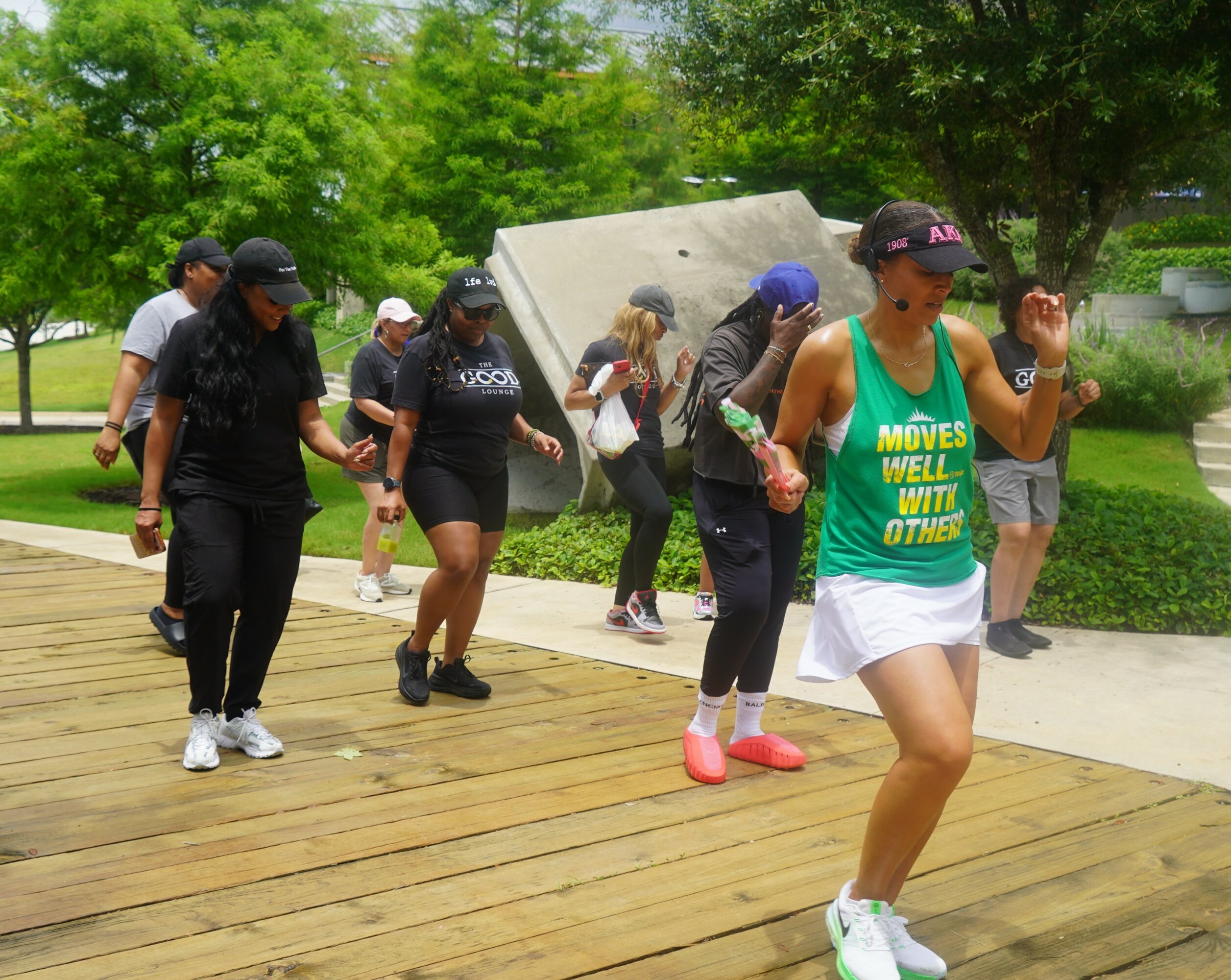Group of people exercising outdoors on a wooden platform, surrounded by green trees and a stone sculpture, with a woman in a green tank top in front.