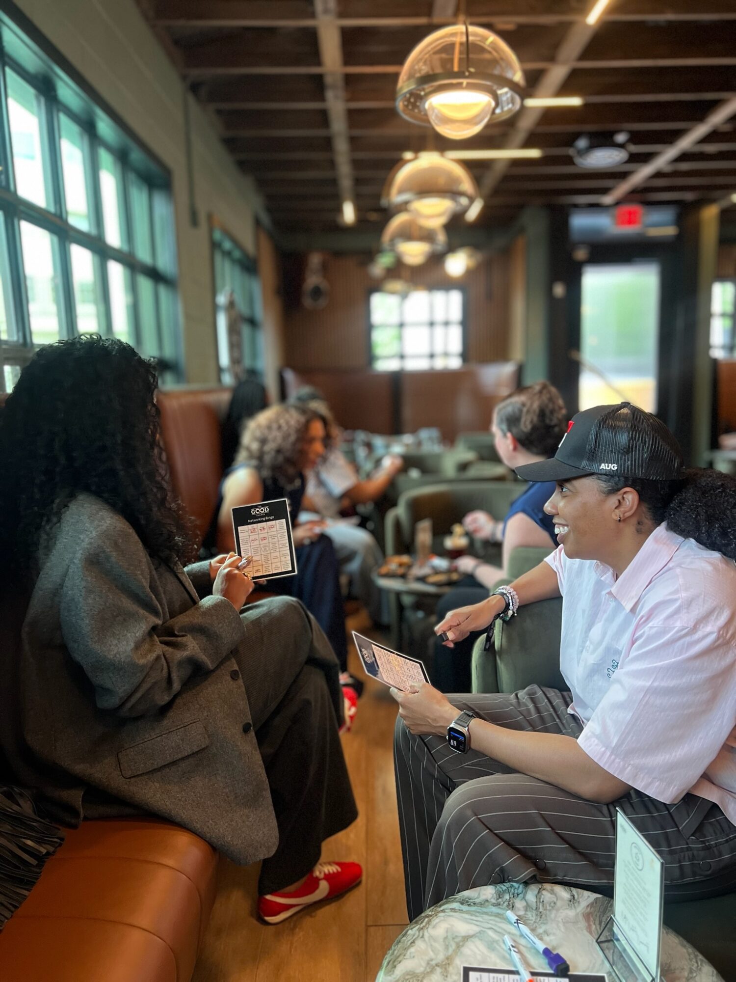 People sitting in a cafe, some using smartphones, with large windows and hanging lights in the background.
