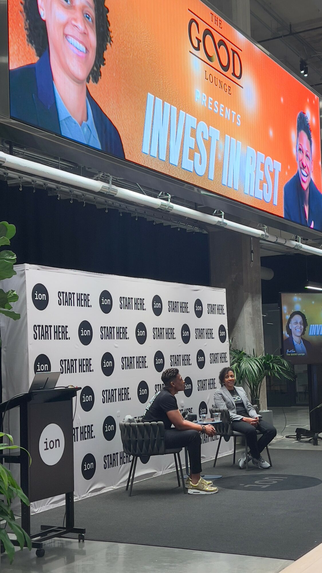 Two people sitting on stage with a backdrop and a large screen above displaying a smiling person and the text 'Invest Invest'.