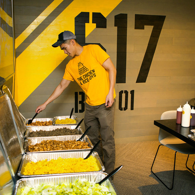 Young man in yellow shirt and cap serving food from buffet trays, with a yellow and black wall behind him.