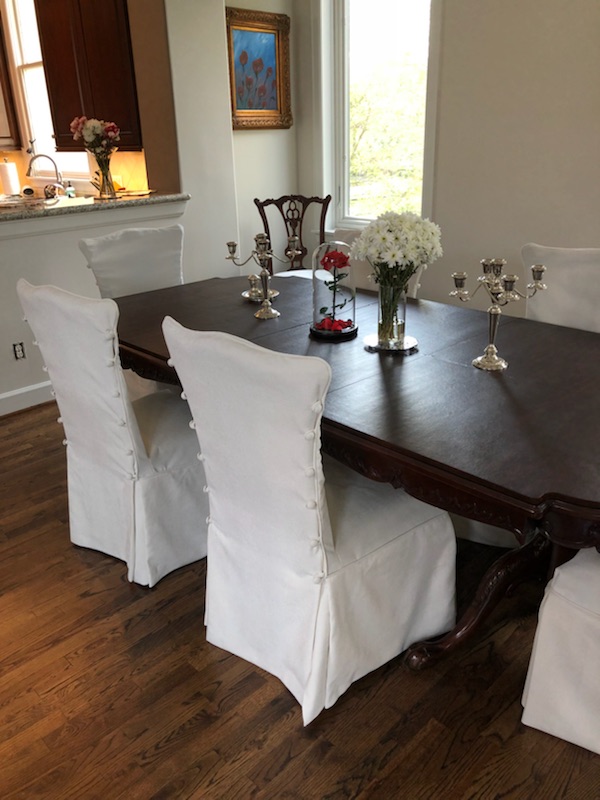 Dining room with dark wooden table, white slipcovered chairs, floral centerpiece, and a window with natural light.