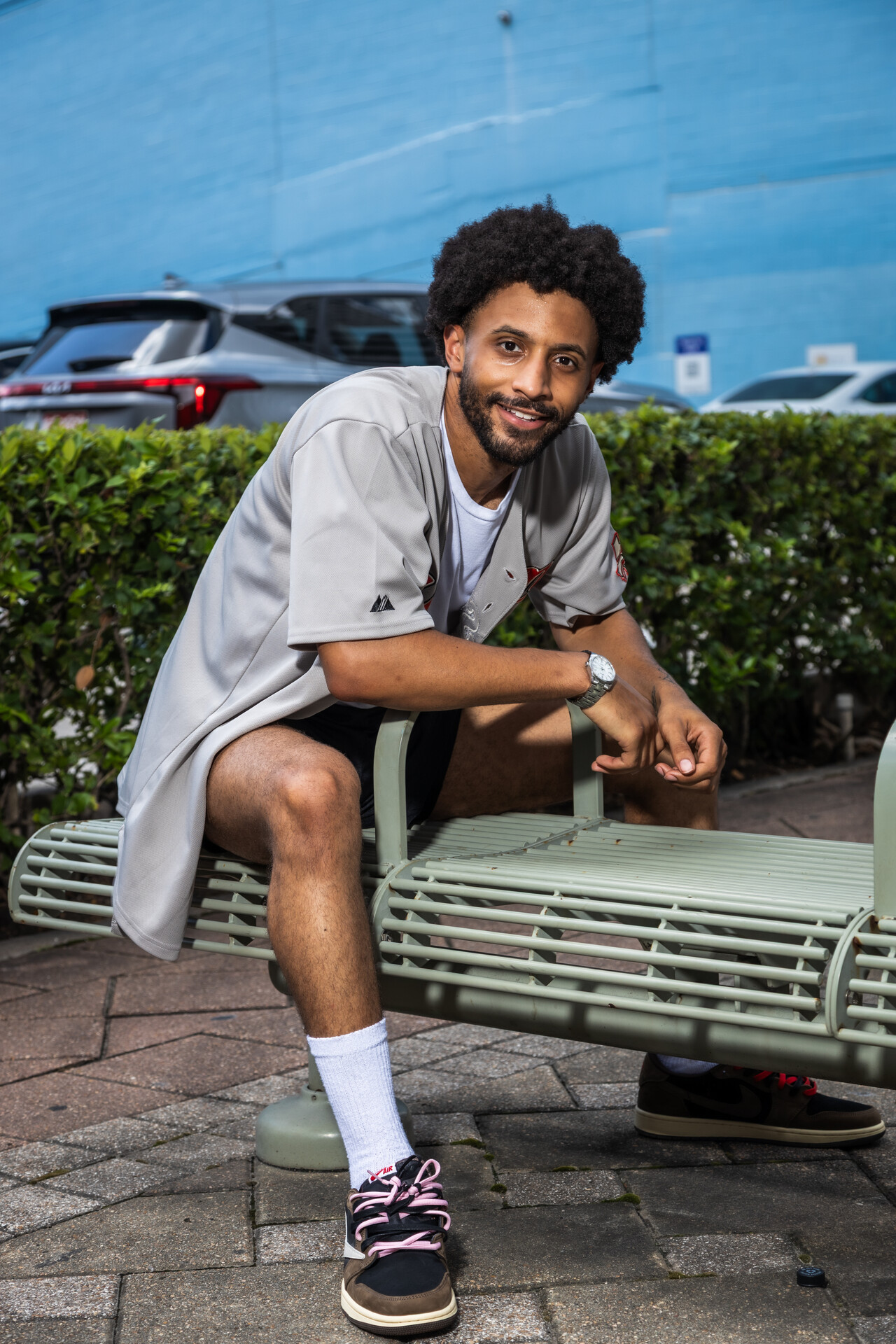 Smiling man with curly hair and beard sitting on a bench outdoors, wearing a light gray shirt, shorts, and sneakers.