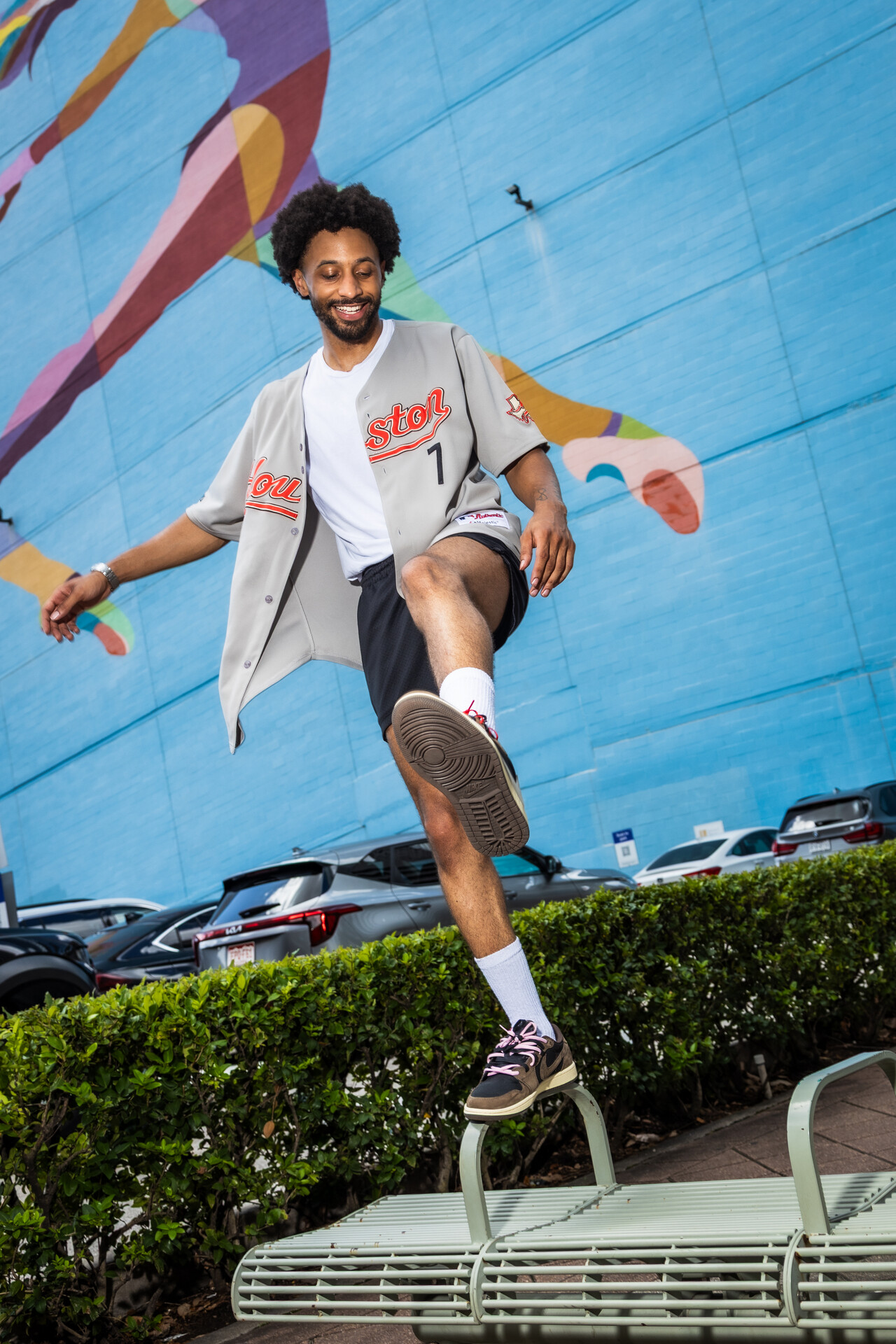 Person with curly hair smiling, balancing on a park bench, wearing a white jacket, black shorts, and sneakers.