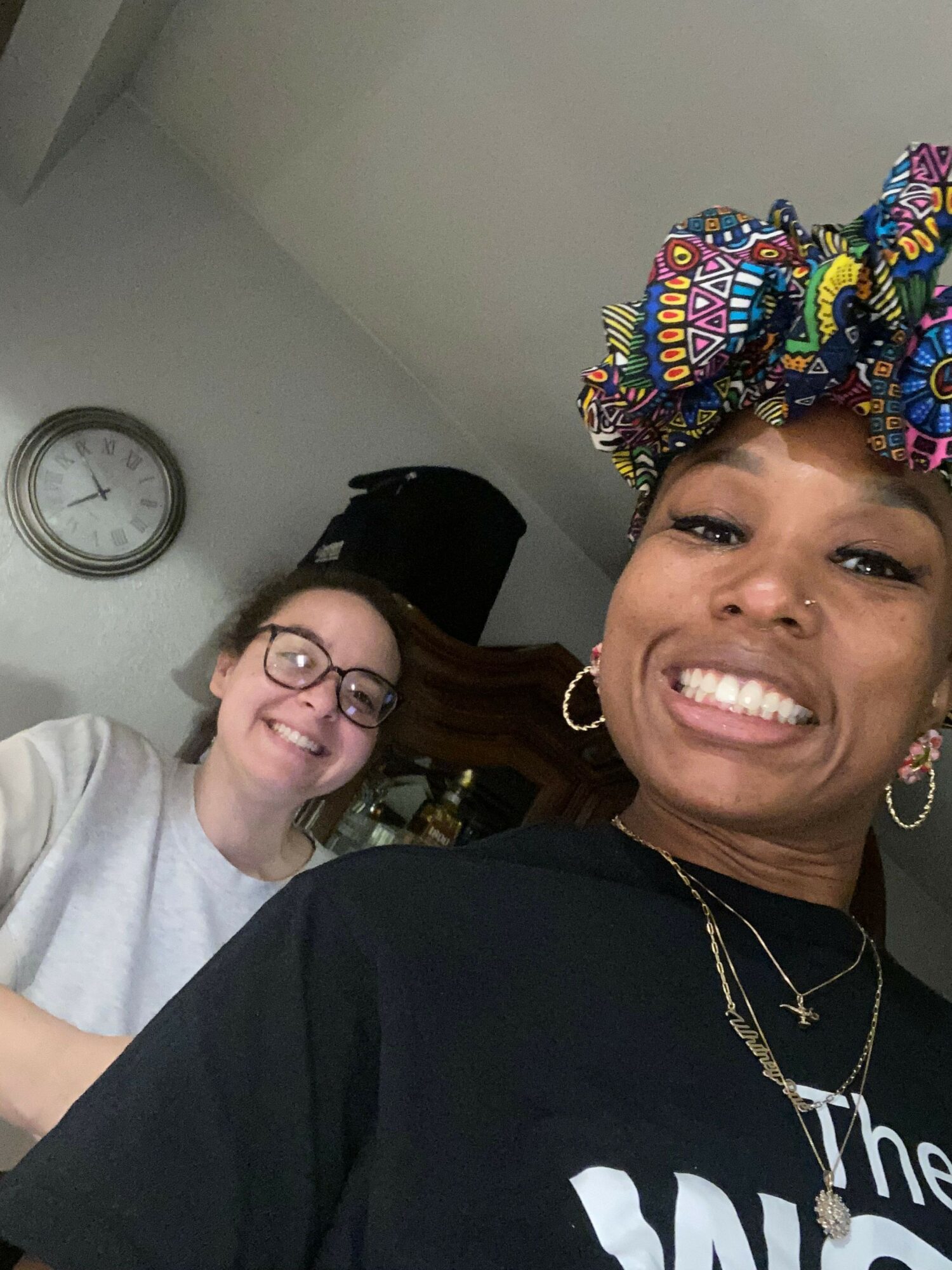Two women smiling, one wearing colorful headwrap, the other with glasses, in an indoor setting with a clock on the wall.