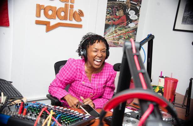 Woman laughing in a radio studio with headphones, microphone, and sound equipment, wearing a pink checkered shirt.