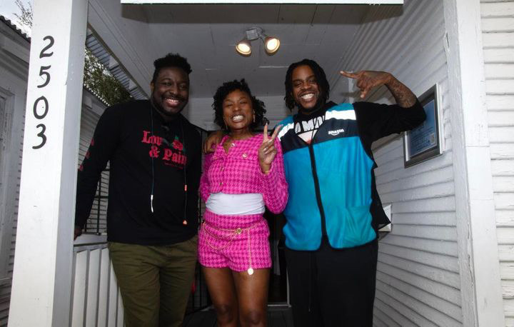 Three people smiling and posing outside a house, with one person making a peace sign, in front of a white wall and door.