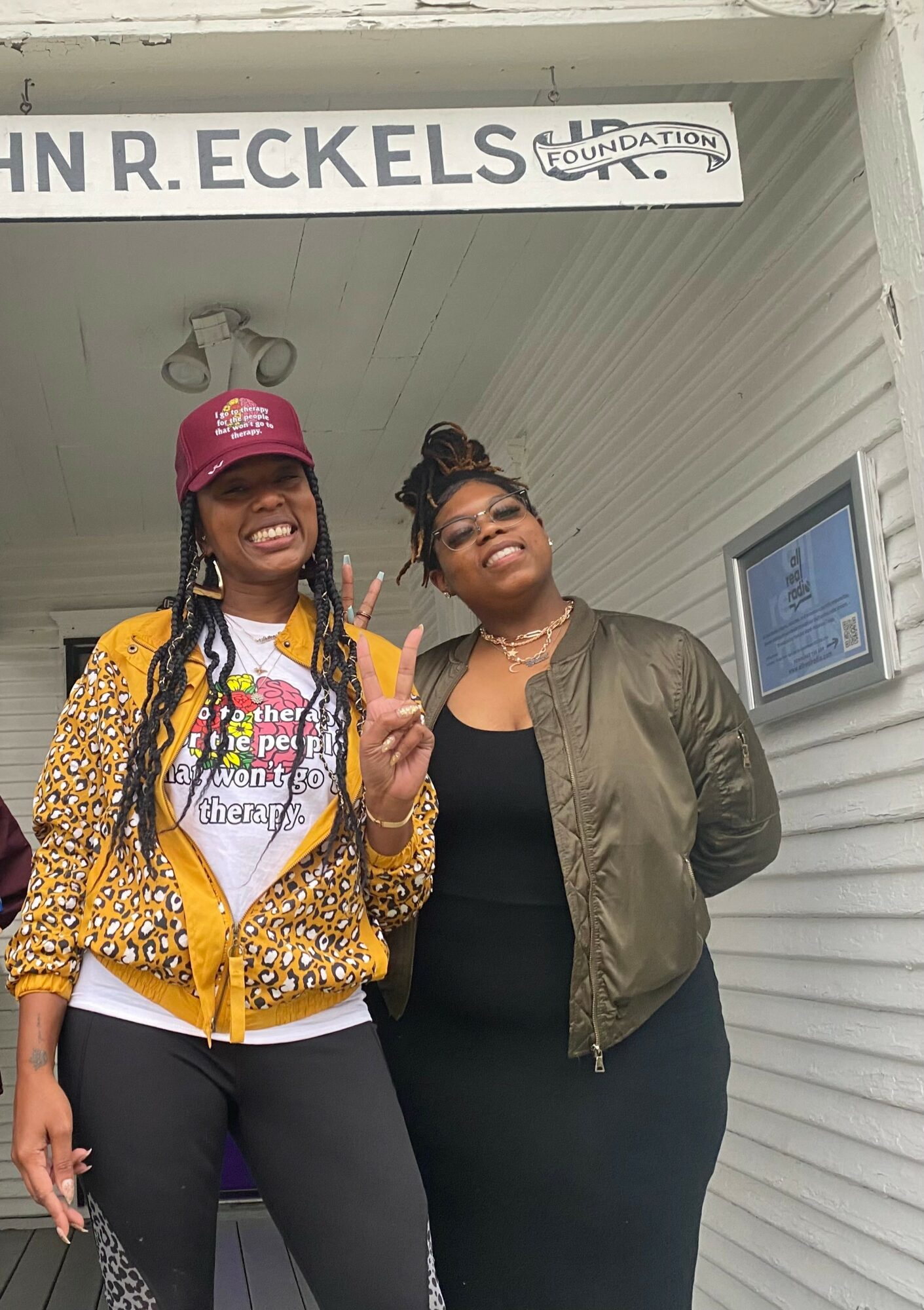 Two women standing outside a building, smiling, one wearing a pink cap and yellow jacket, the other in a black outfit and glasses.
