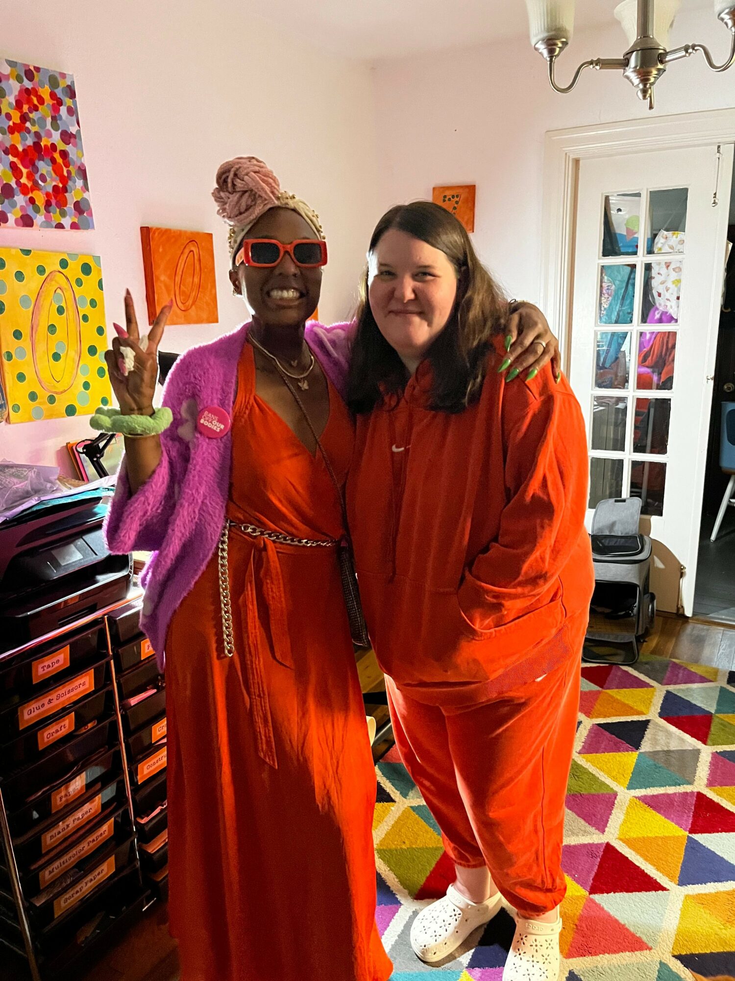 Two women in colorful outfits pose indoors, one making a peace sign, with a vibrant geometric rug and artwork in background.