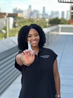 Smiling woman holding a small object towards the camera on a rooftop with cityscape background.