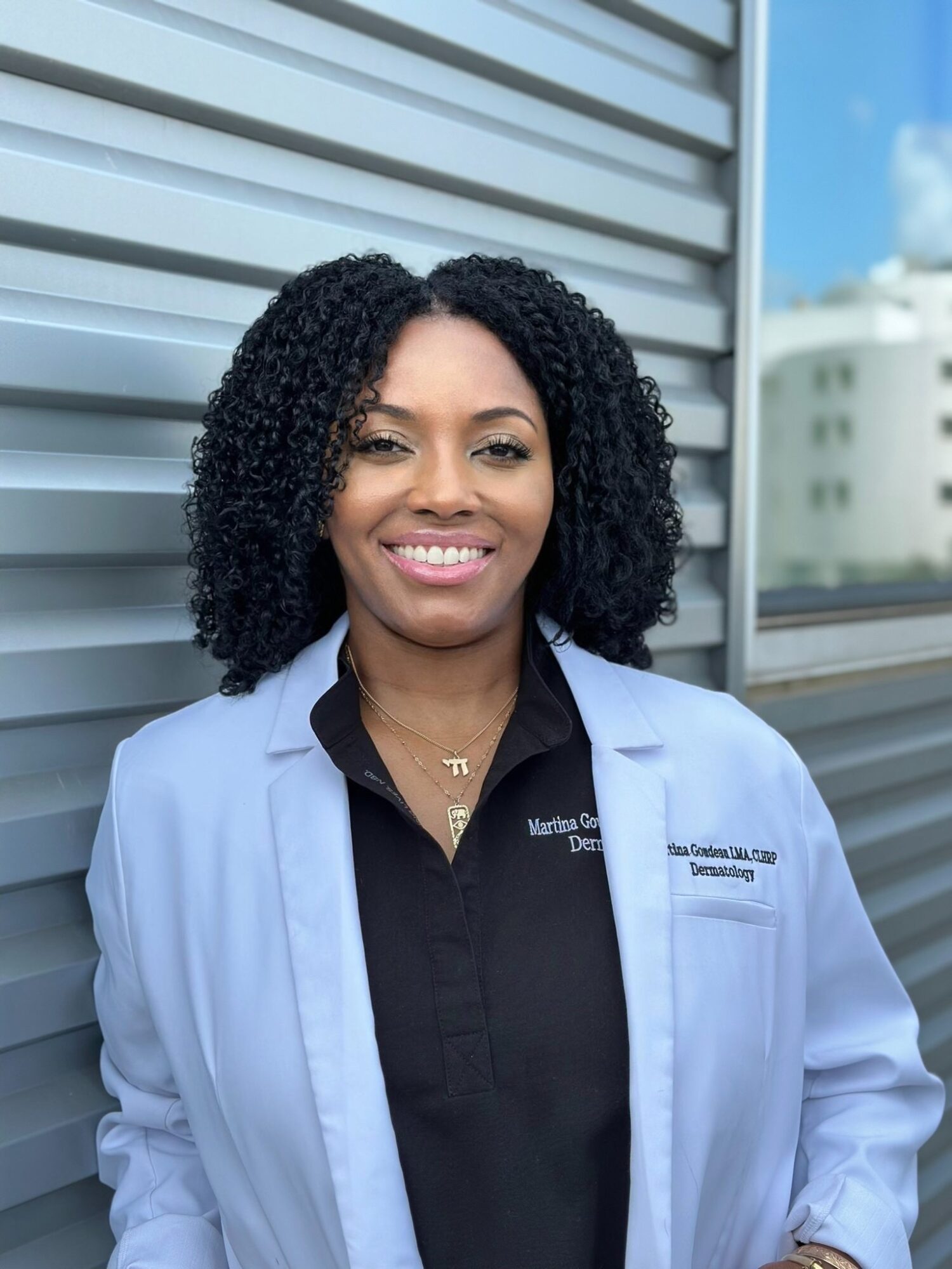 Smiling woman with curly hair in a light blue lab coat outdoors, with a building and sky in background.