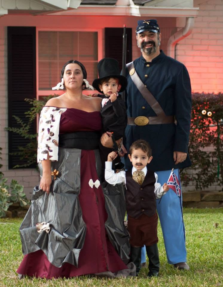 Family dressed in historical costumes standing outside a house with a garden, smiling at the camera.