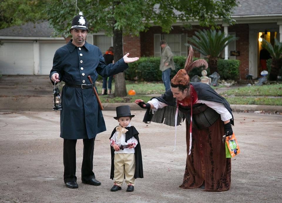 Two adults and a child in costumes on a street, with houses and trees in the background.