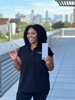 Smiling woman holding a small object and a paper on a rooftop with city skyline in background.