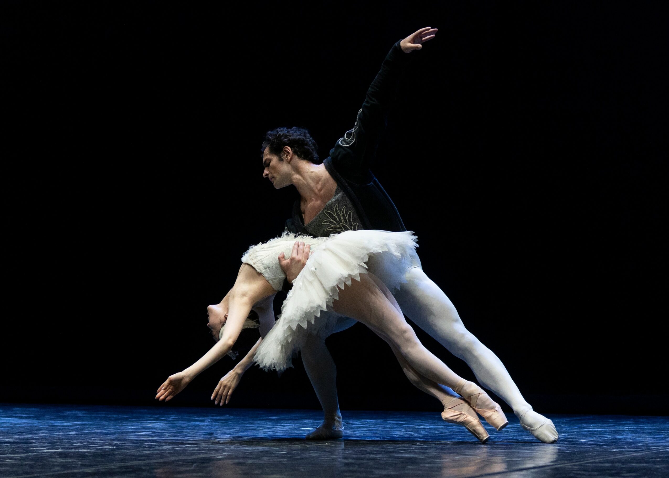 Two ballet dancers perform on stage, one supporting the other in a pose, with a dark background.