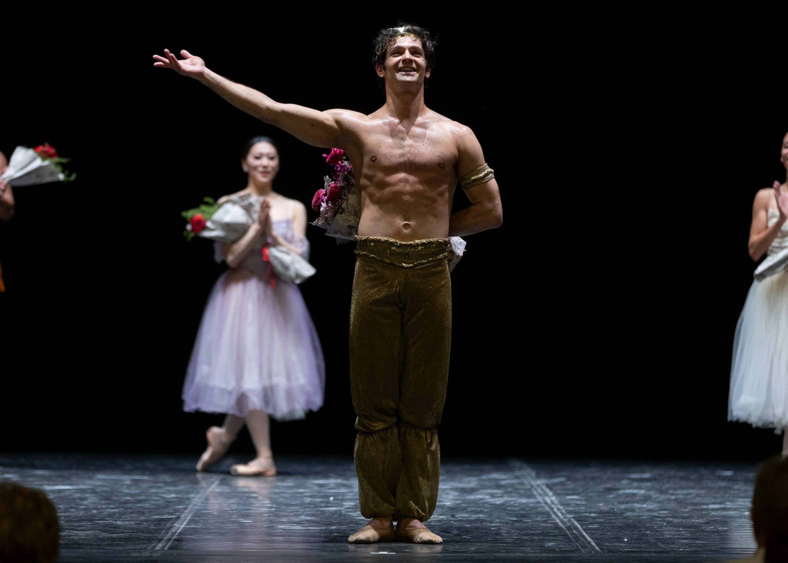 Man standing on stage with arms outstretched, smiling, surrounded by women in ballet costumes holding flowers.