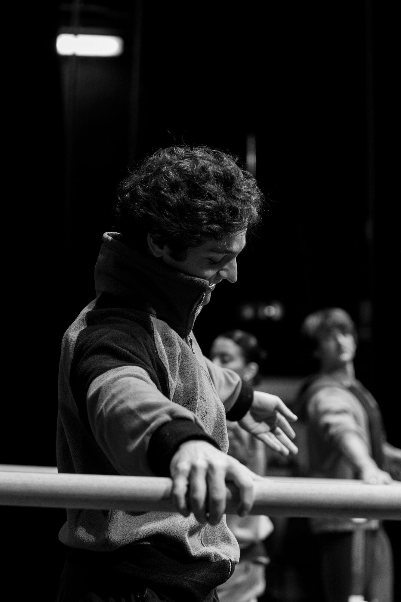 Person with curly hair leaning on a railing, with other people in the background, in a dark setting.