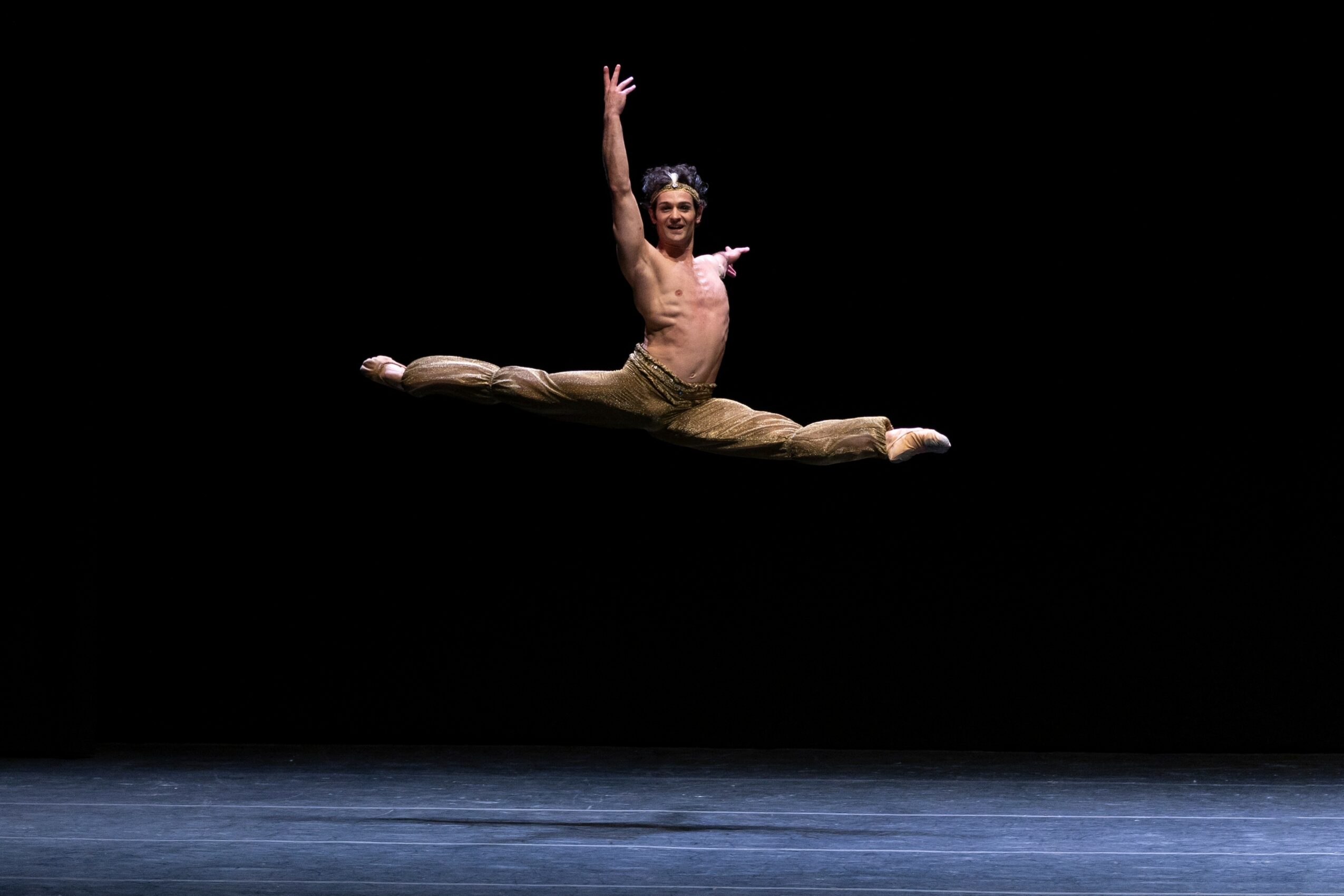 A male dancer performs a split leap mid-air against a black background, with arms raised and legs extended sideways.