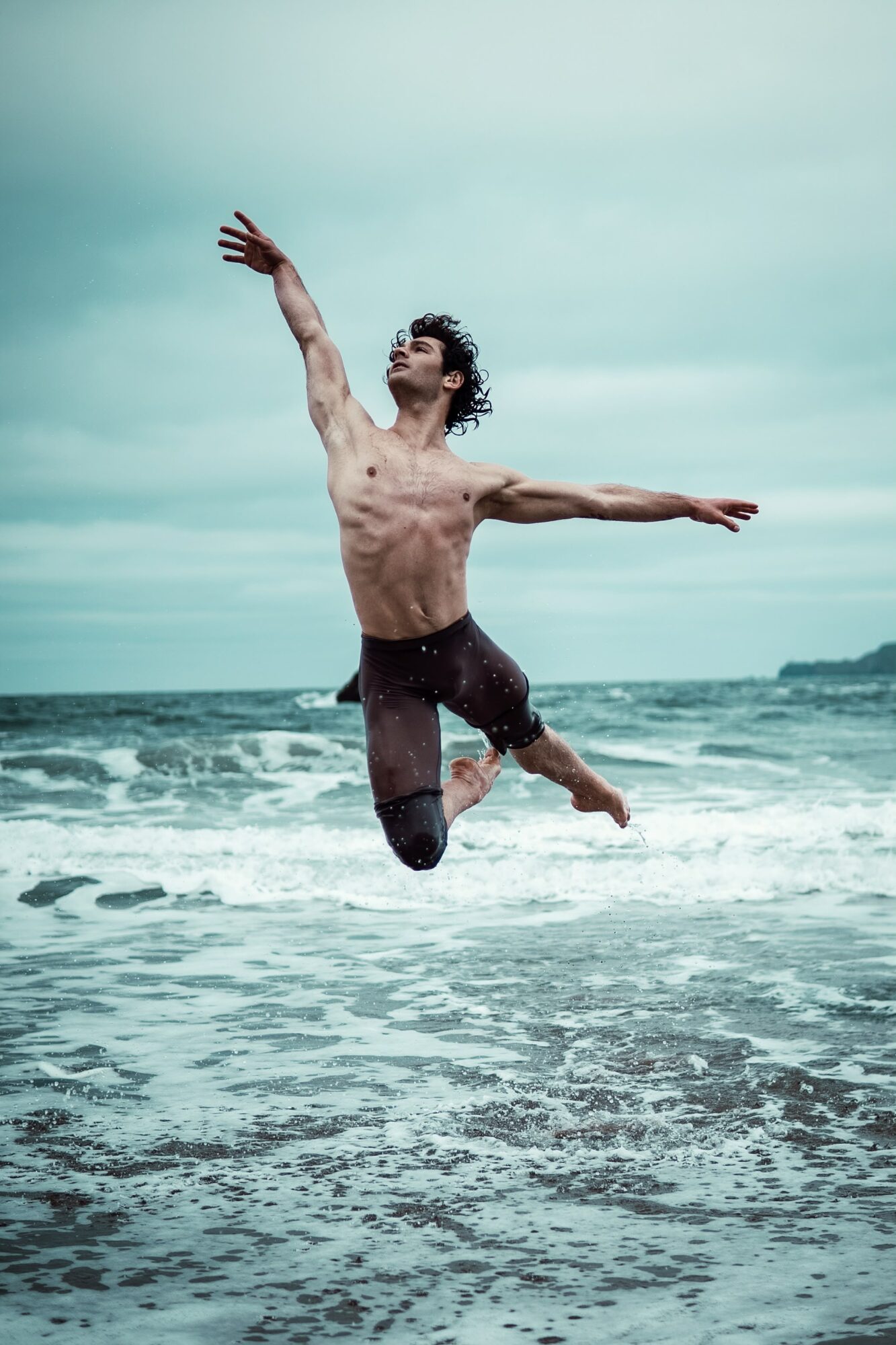 Young man jumping in the ocean with arms outstretched, waves around him, overcast sky in background.