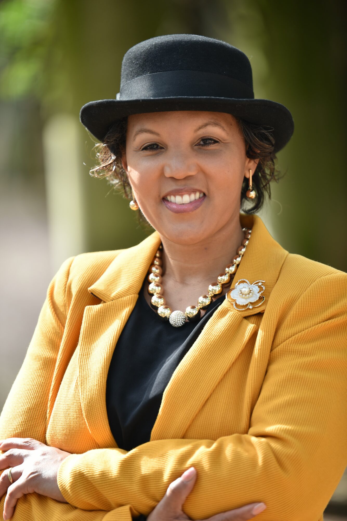 Woman wearing a black hat, yellow blazer, pearl necklace, and earrings outdoors, smiling with arms crossed.