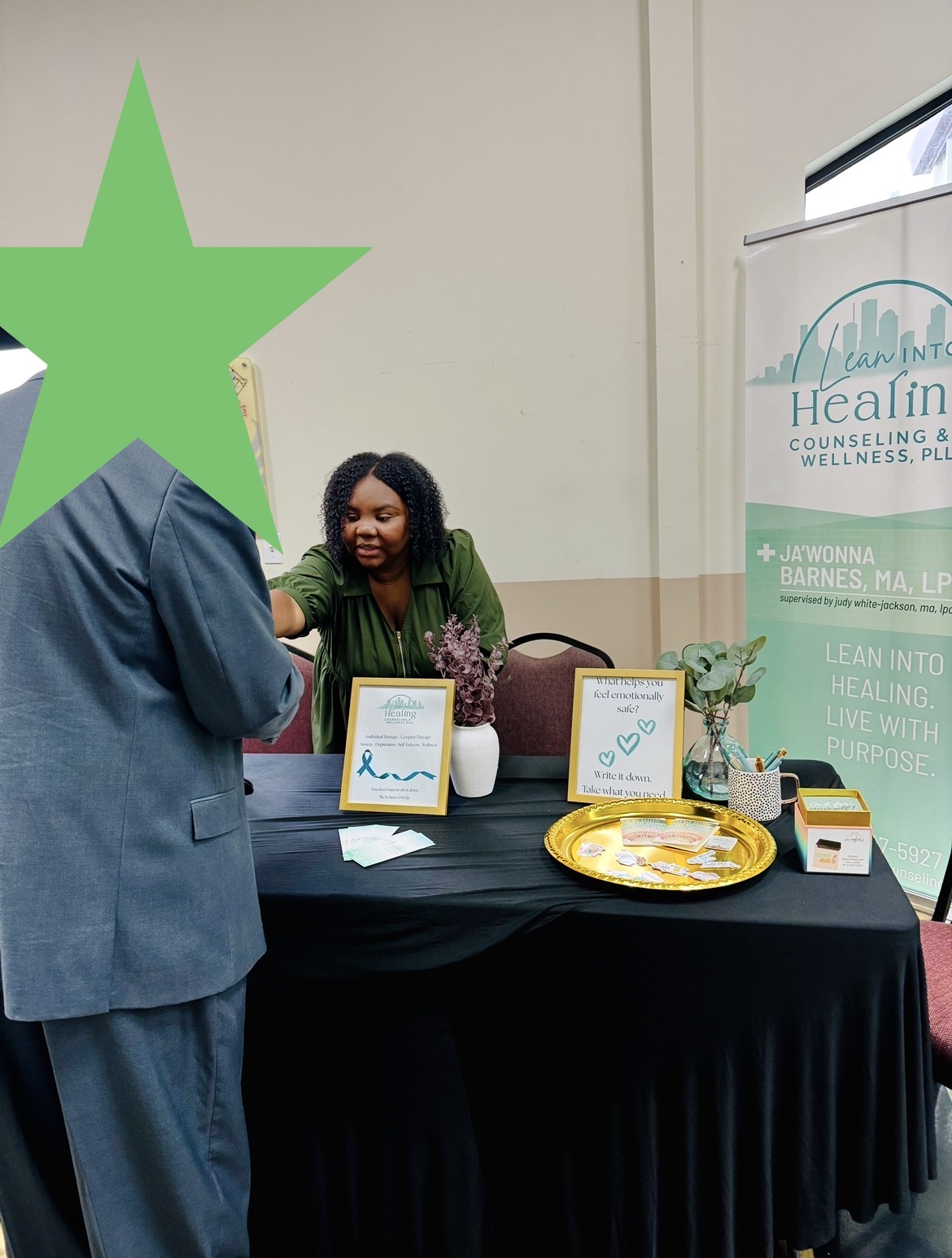 Woman standing behind a table with promotional materials, smiling, at an indoor event. A large green star overlays part of the image.