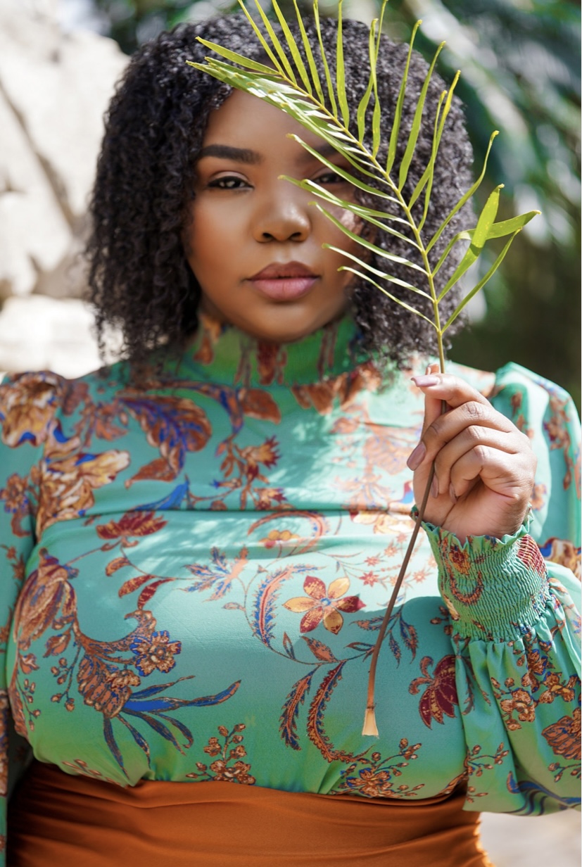 Woman with curly hair holding a palm leaf in front of her face, wearing a colorful patterned top.