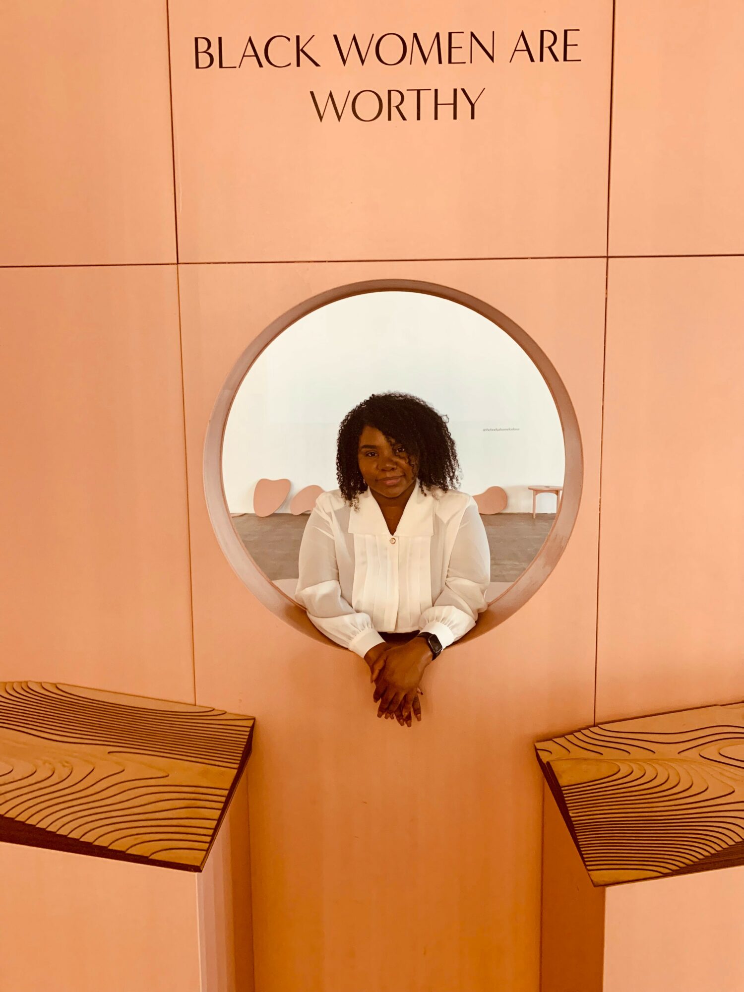 Woman with curly hair in white shirt looking through a circular window, pink wall with text above, two wooden benches below.