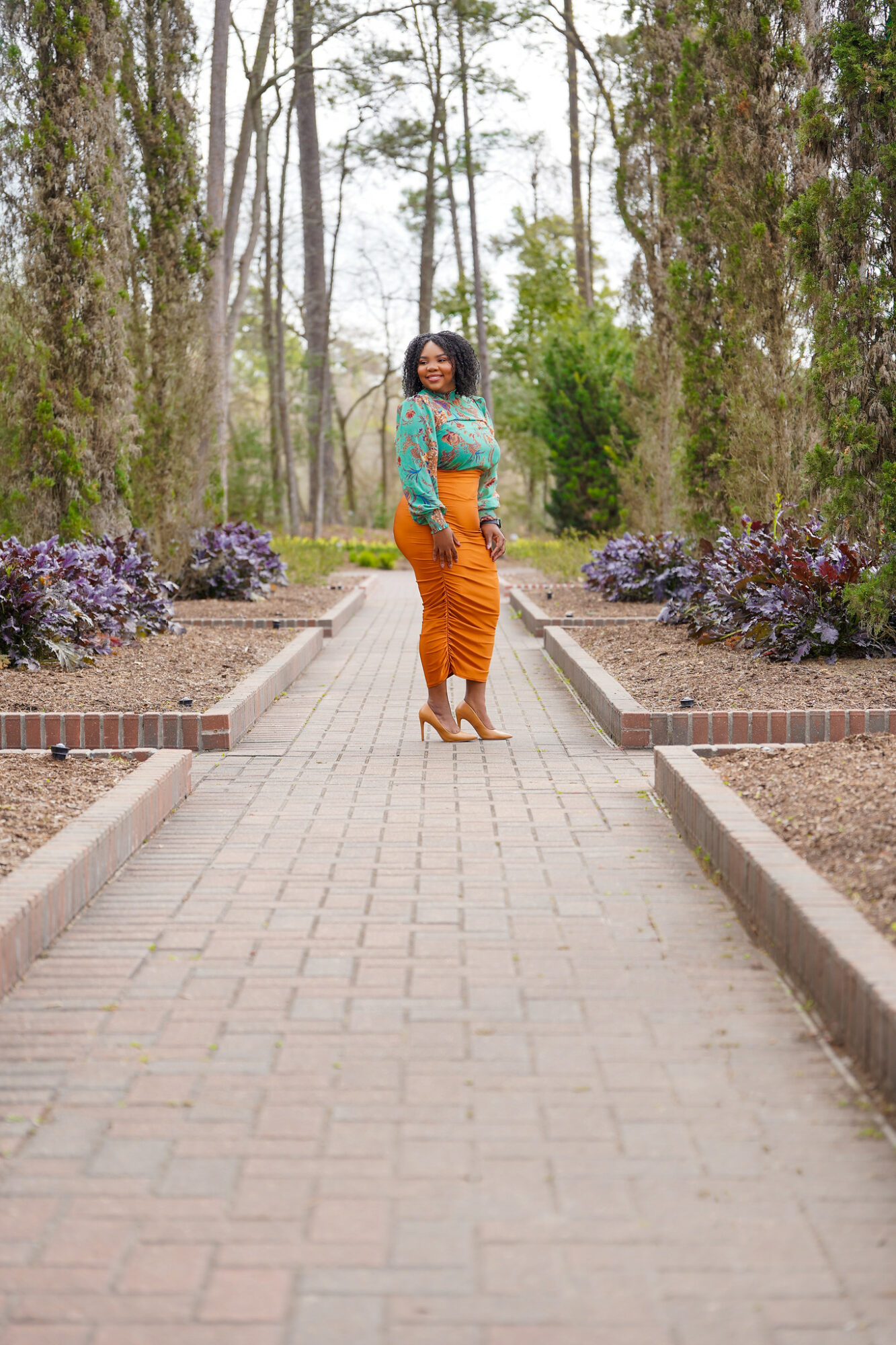 Woman standing on a brick pathway in a park with trees and purple flowers, smiling, wearing a teal top and orange skirt.