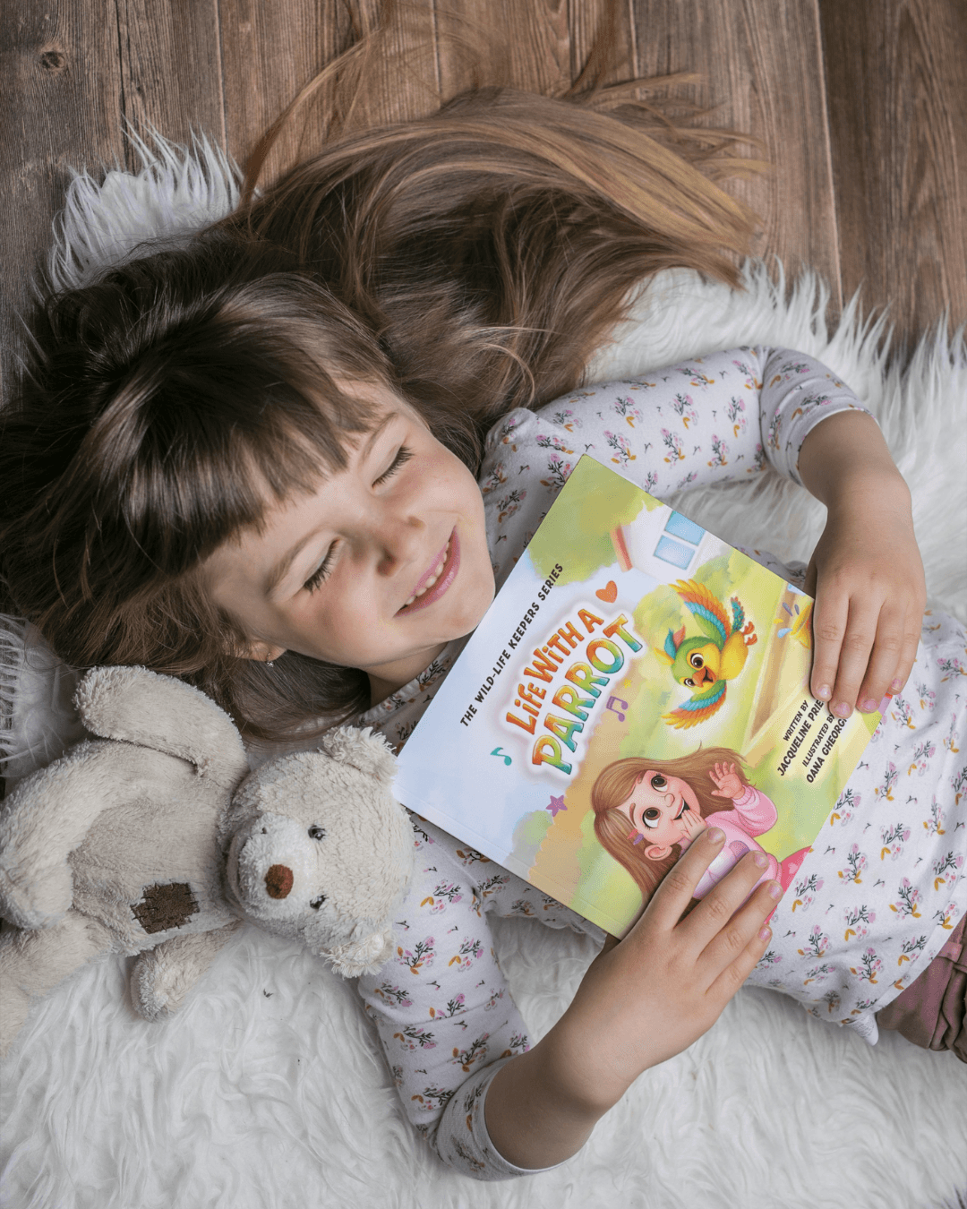 Girl lying on a white furry surface, holding a colorful children's book, with stuffed animals nearby, smiling with eyes closed.
