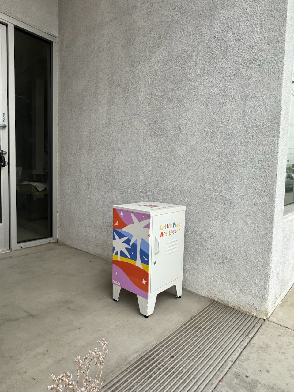 White box with colorful star and rainbow design on a concrete sidewalk, next to a wall and a drain grate.