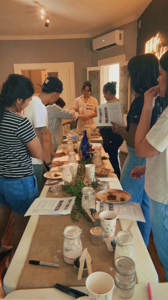 People gathered around a table with food and drinks, reading papers, in a room with a wall-mounted air conditioner.