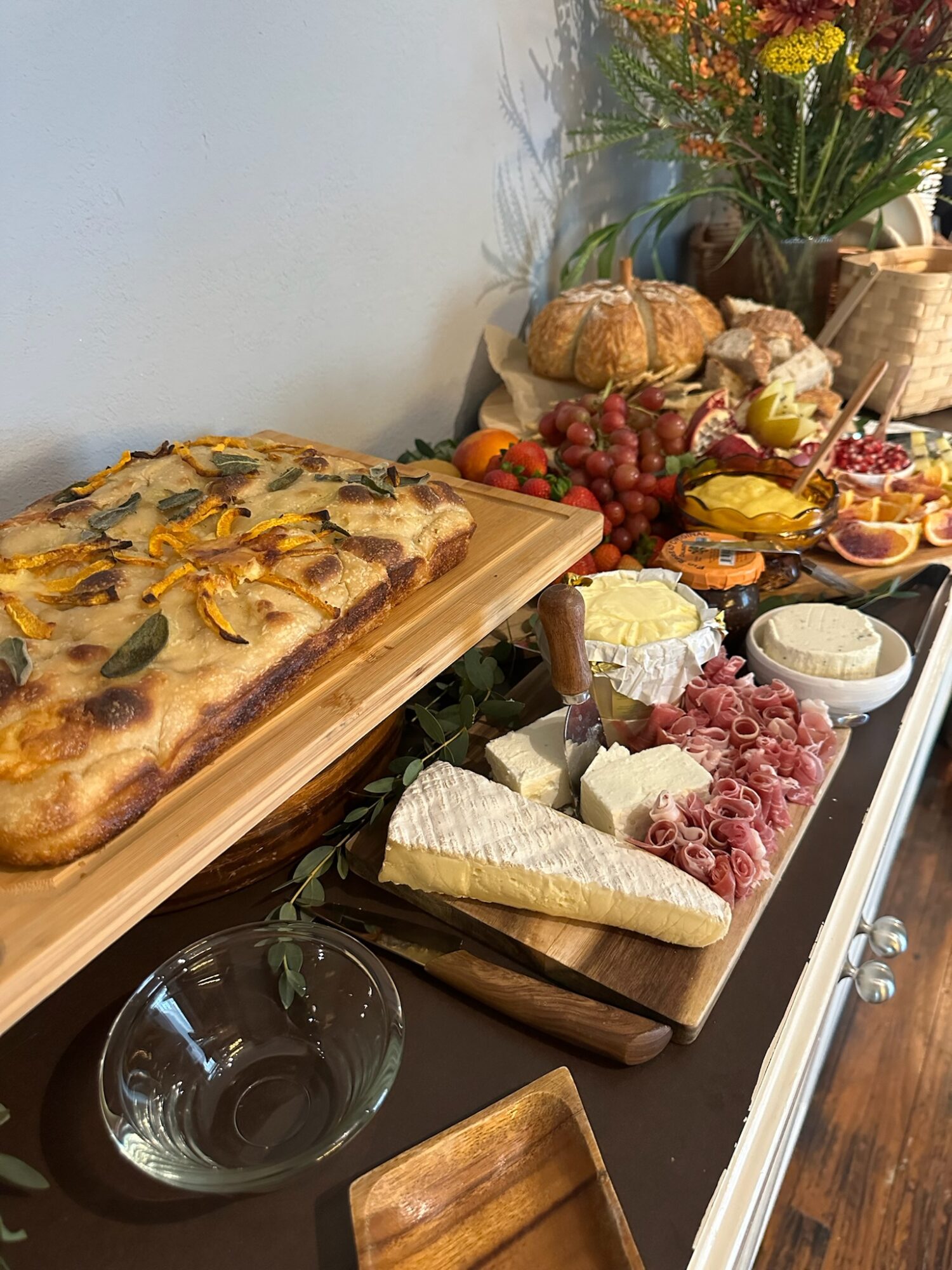 A spread of food including a large rectangular pizza, cheese, meats, grapes, bread, and flowers on a table.