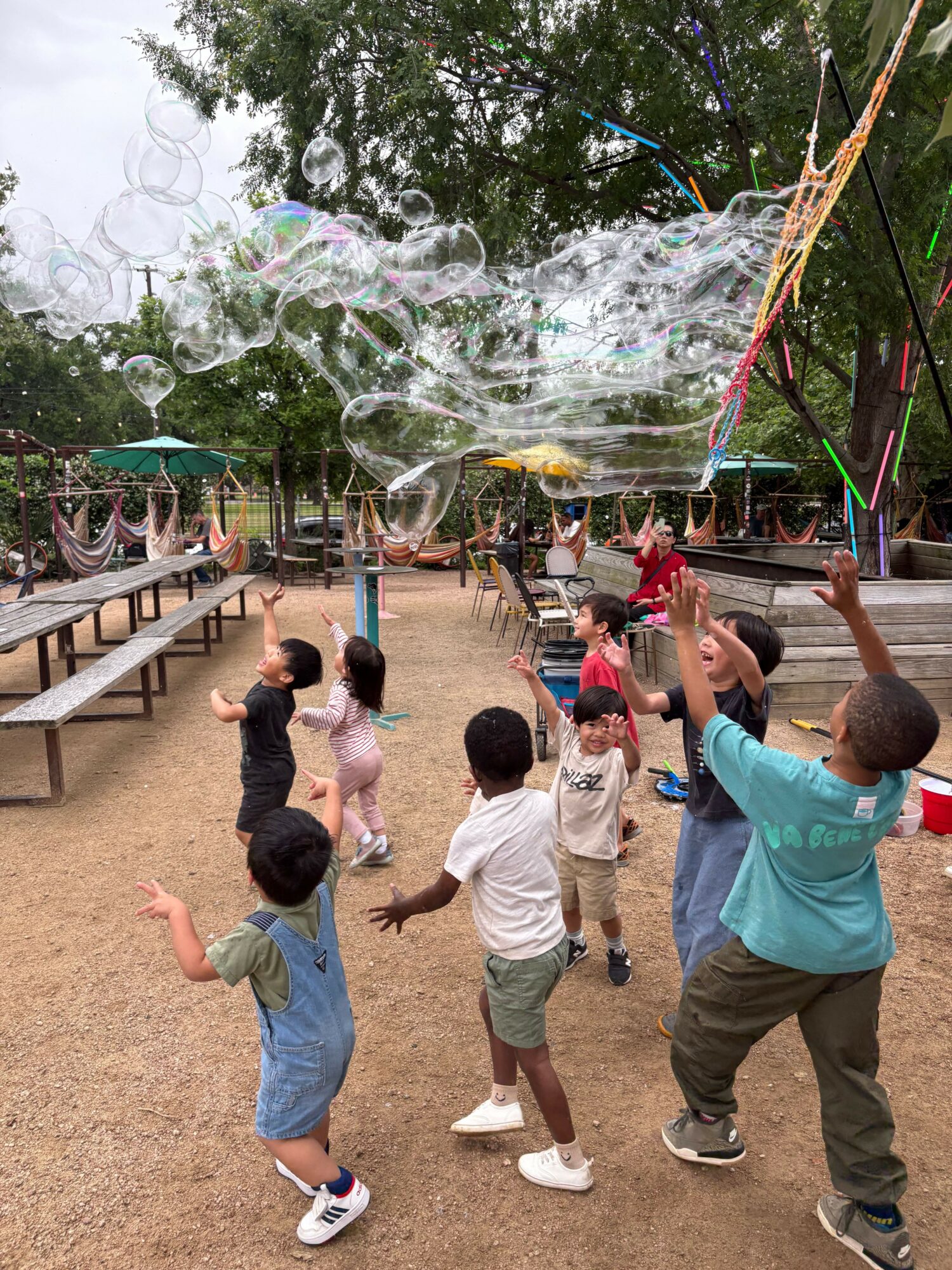 Children playing outdoors with bubbles and colorful decorations, some reaching for bubbles, on a sandy ground with trees and structures.