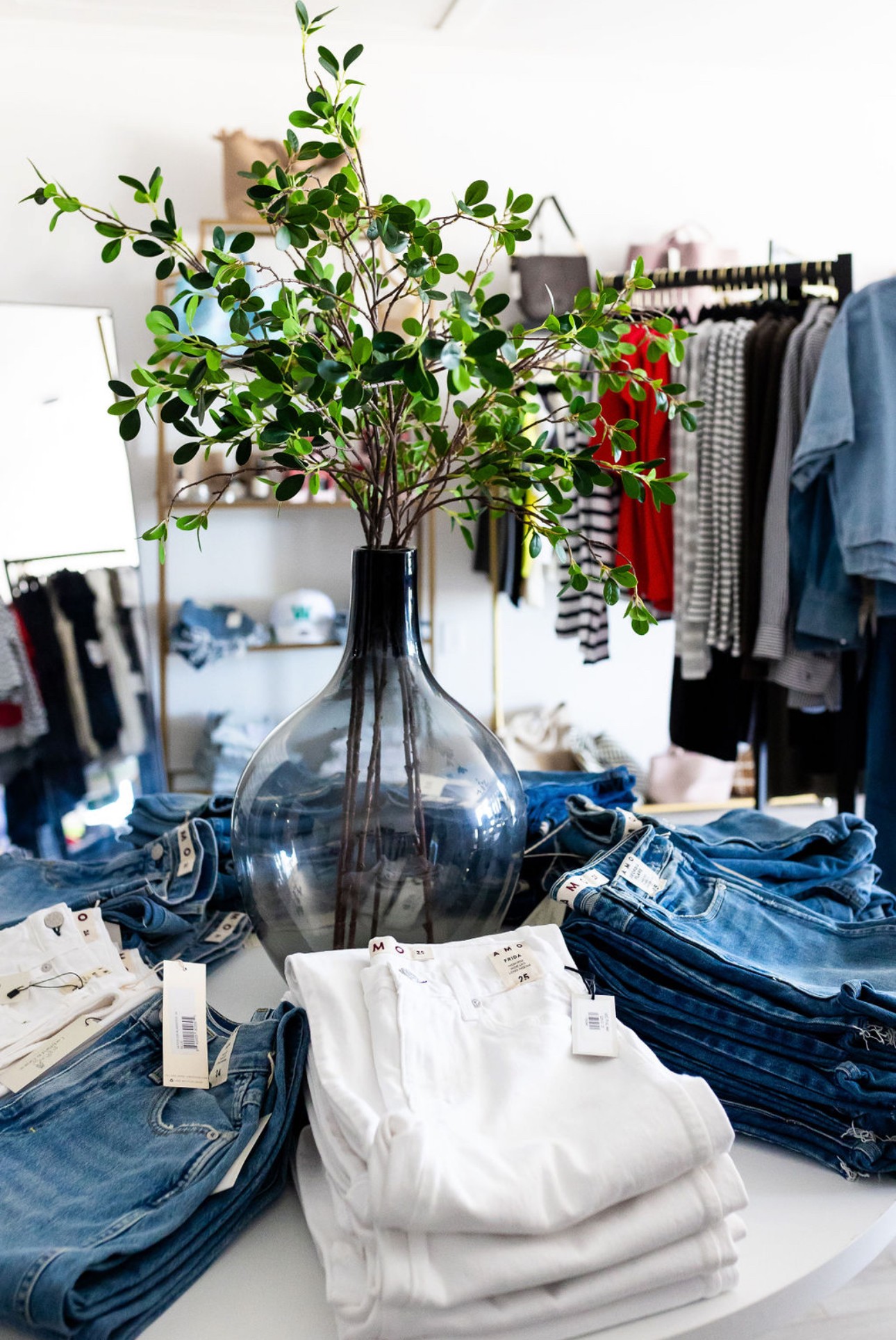 A large glass vase with green leafy branches on a table surrounded by folded jeans and clothing in a store.