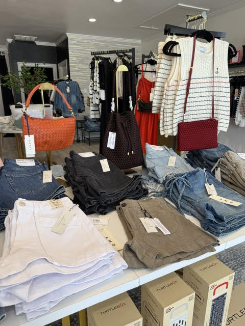 Display of handbags, folded clothes, and boxes in a retail store with clothing racks in the background.