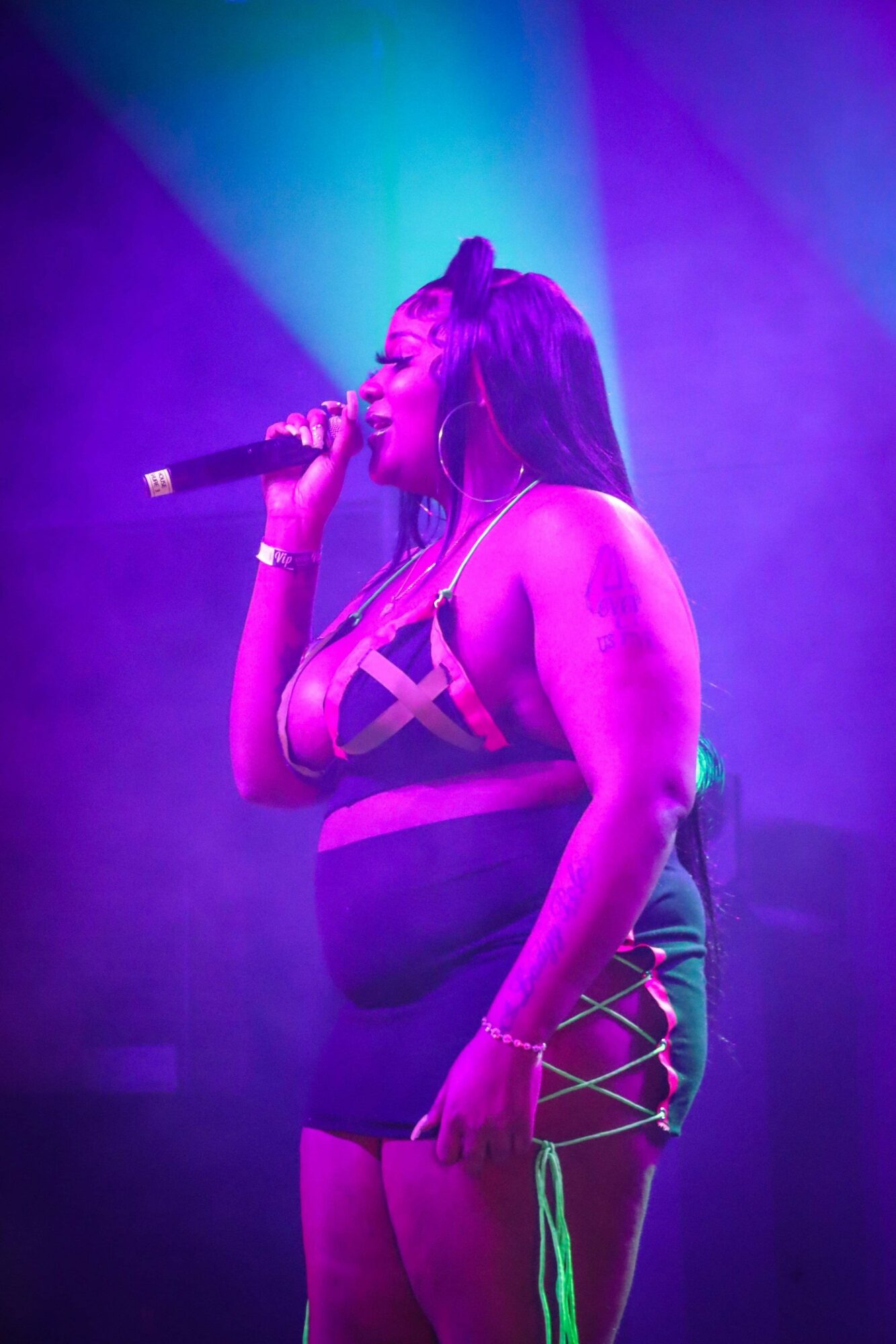 Performer singing into a microphone on stage with colorful purple and blue lighting, wearing a strappy outfit.