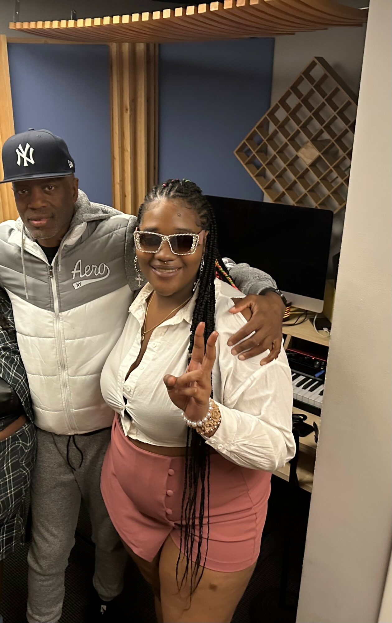 Two people in a recording studio, one woman smiling and making a peace sign, the other person partially visible.