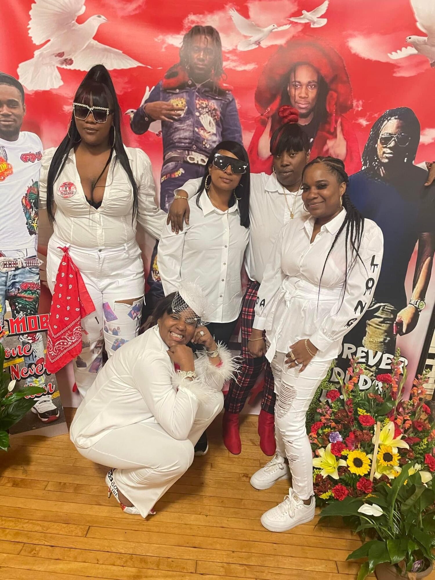 Group of seven people posing indoors with a red and white background, some wearing white outfits and sunglasses, surrounded by flowers.