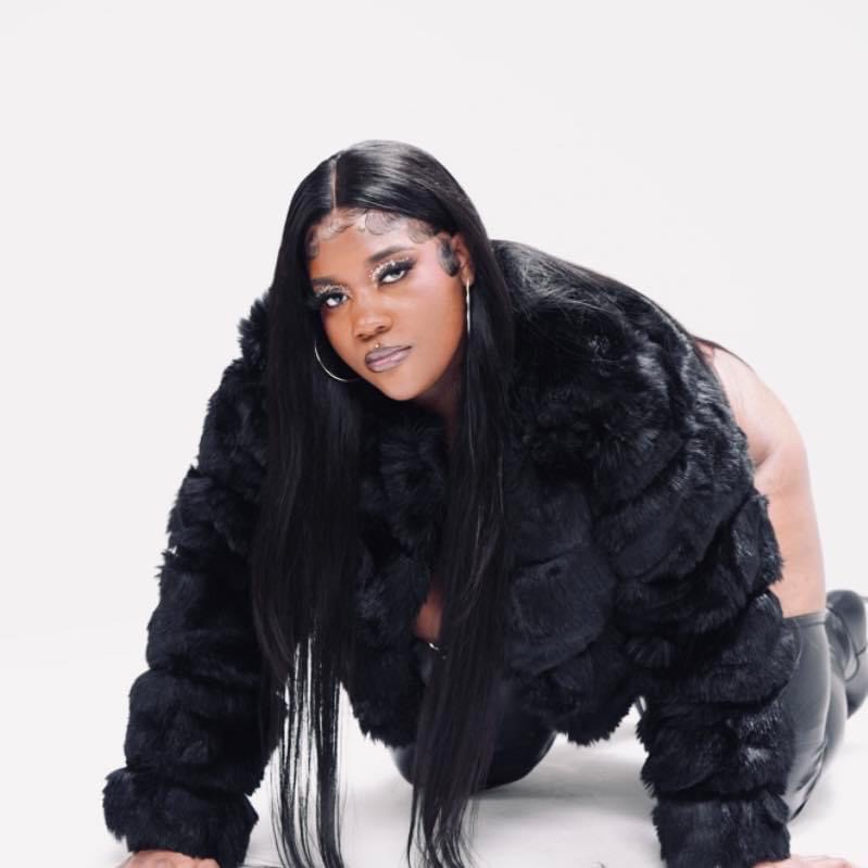 Woman with long black hair wearing a black fur coat, crouching on the floor against a white background.