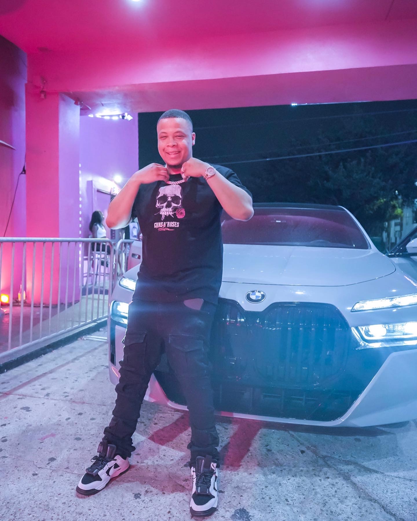 Young man standing in front of a car under a pink-lit structure, smiling and making a gesture with his hands.