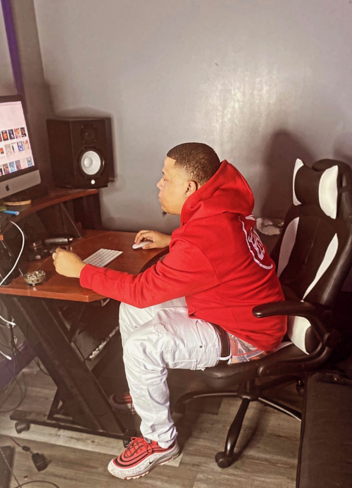 Young person sitting at a desk using a computer, wearing a red hoodie and white pants, in a room with a speaker and chair.