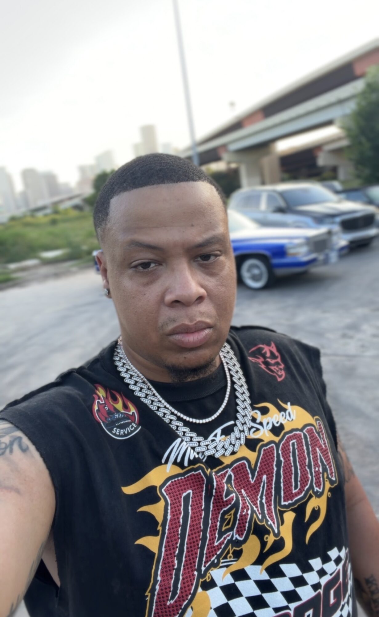 Young man with short hair and jewelry taking a selfie outdoors with cars and buildings in background.