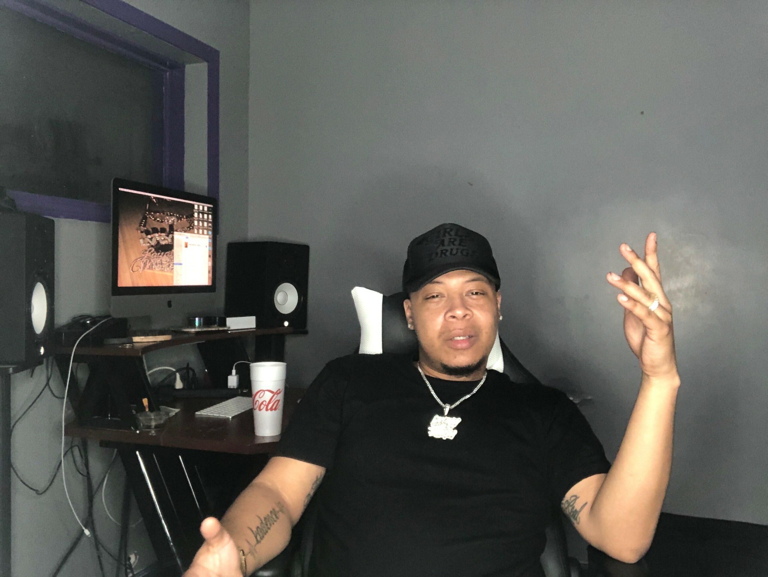 Person sitting in a room with a computer, speakers, and a drink on the desk, making a hand gesture.