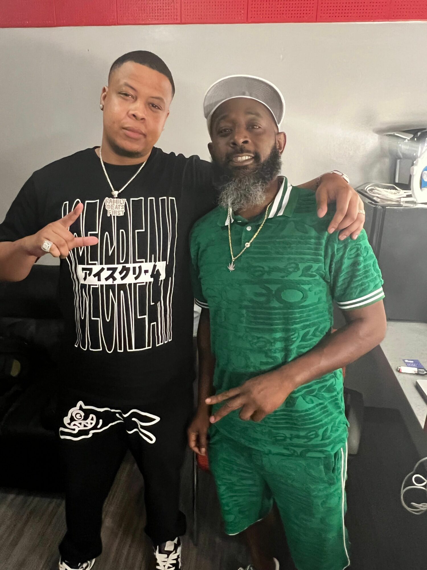 Two men standing indoors, one in a green outfit and the other in a black hoodie, posing for a photo.