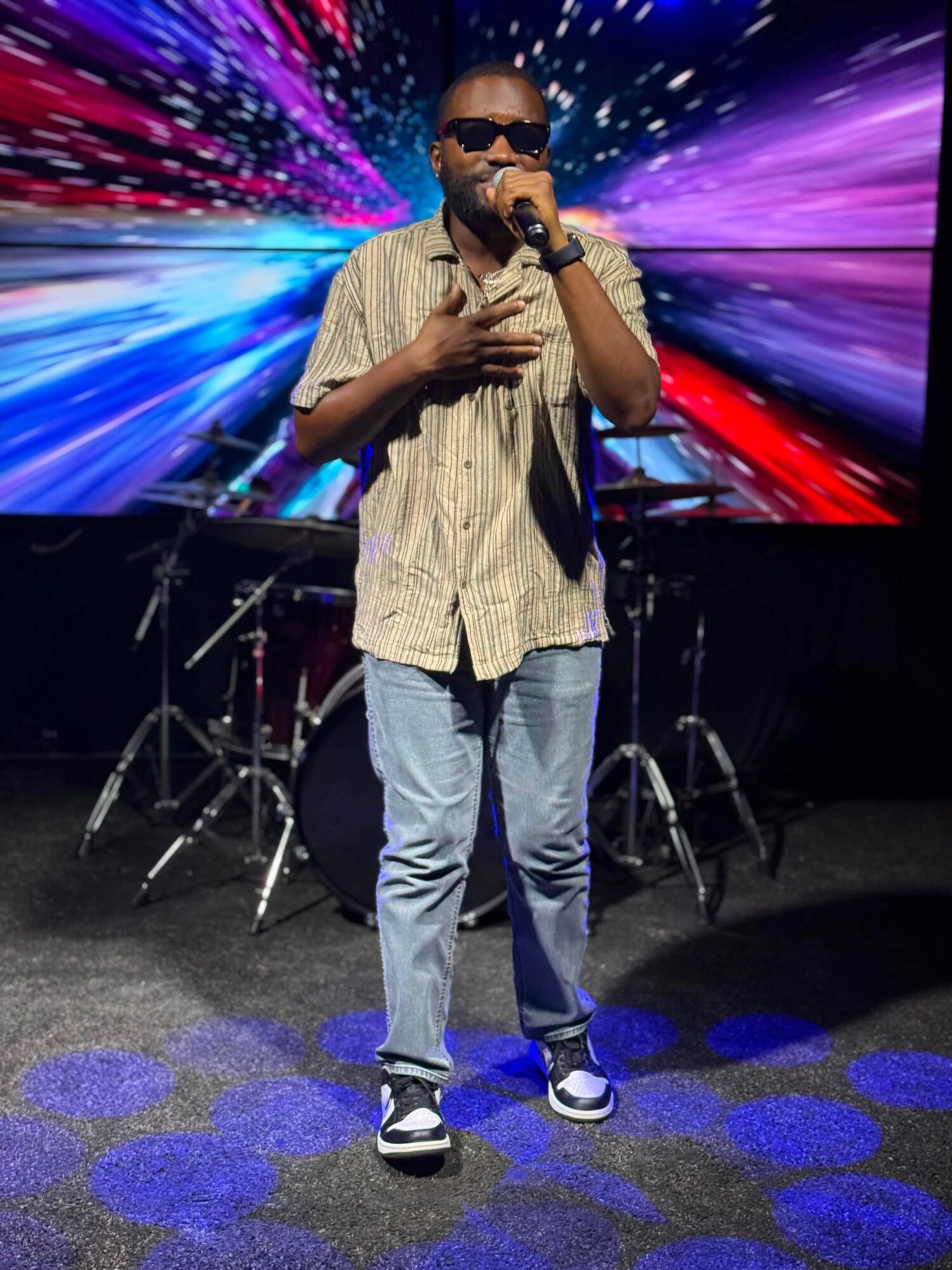 Man singing into microphone on stage with colorful digital background, drums behind him, wearing sunglasses, striped shirt, jeans, and sneakers.
